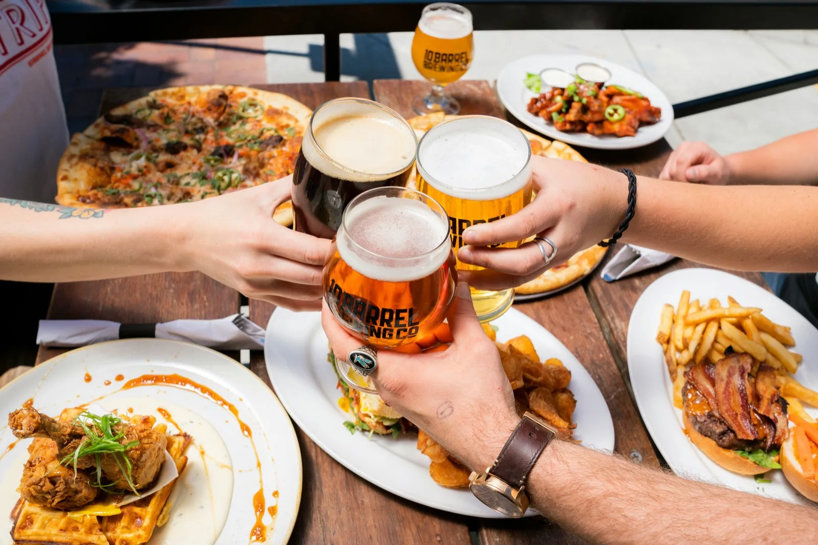 Friends clinking beer glasses over pub food on a sunny outdoor terrace