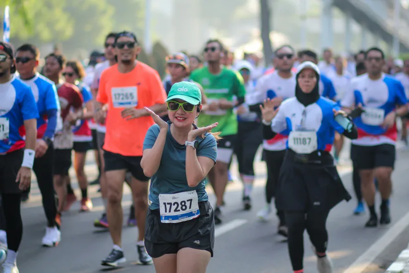 A runner mid-race smiles at the camera in a crowd of fun-run participants, bib numbers visible, on a bright sunny day